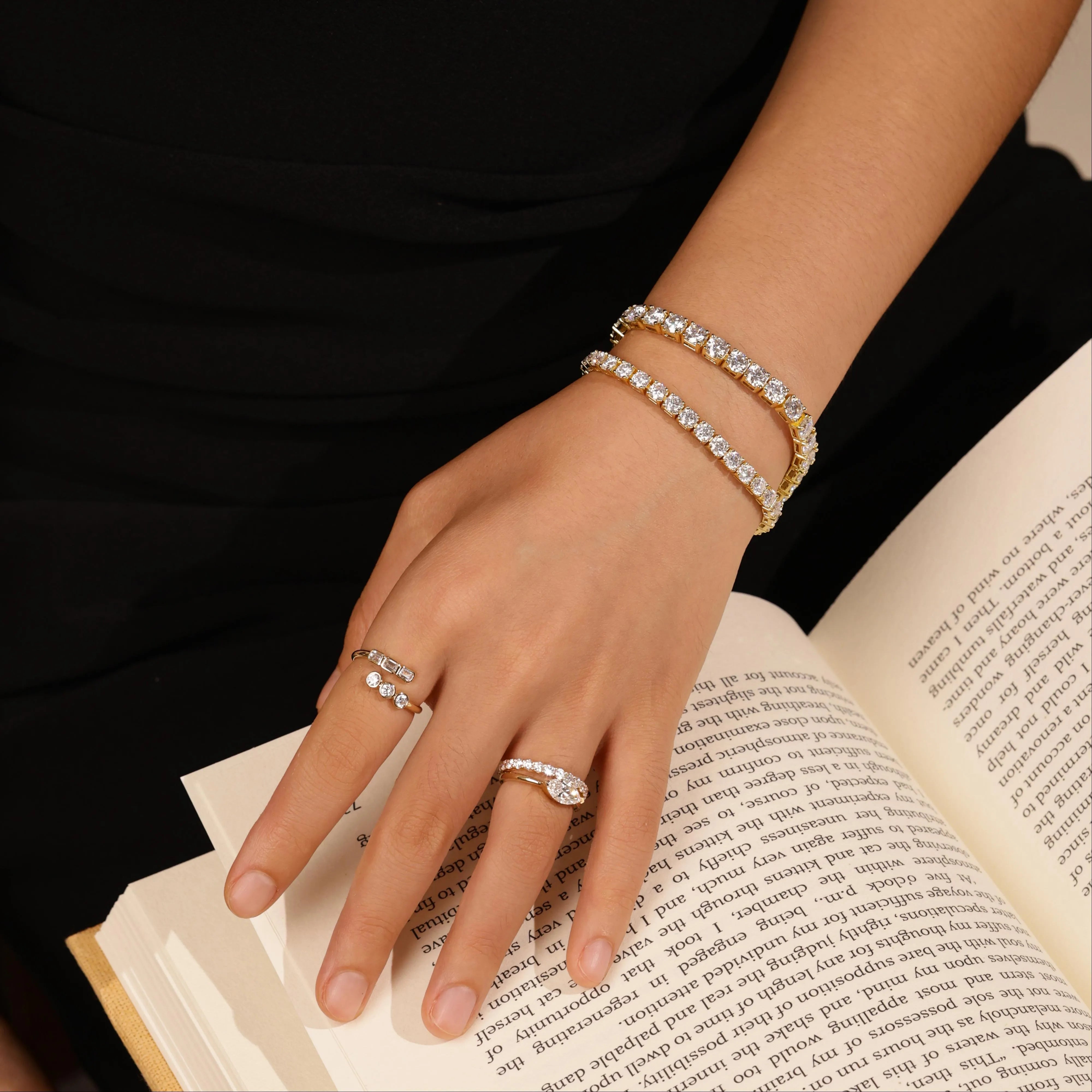 Woman hands resting on open book, wearing stacked gold cubic zirconia tennis bracelets and diamond rings