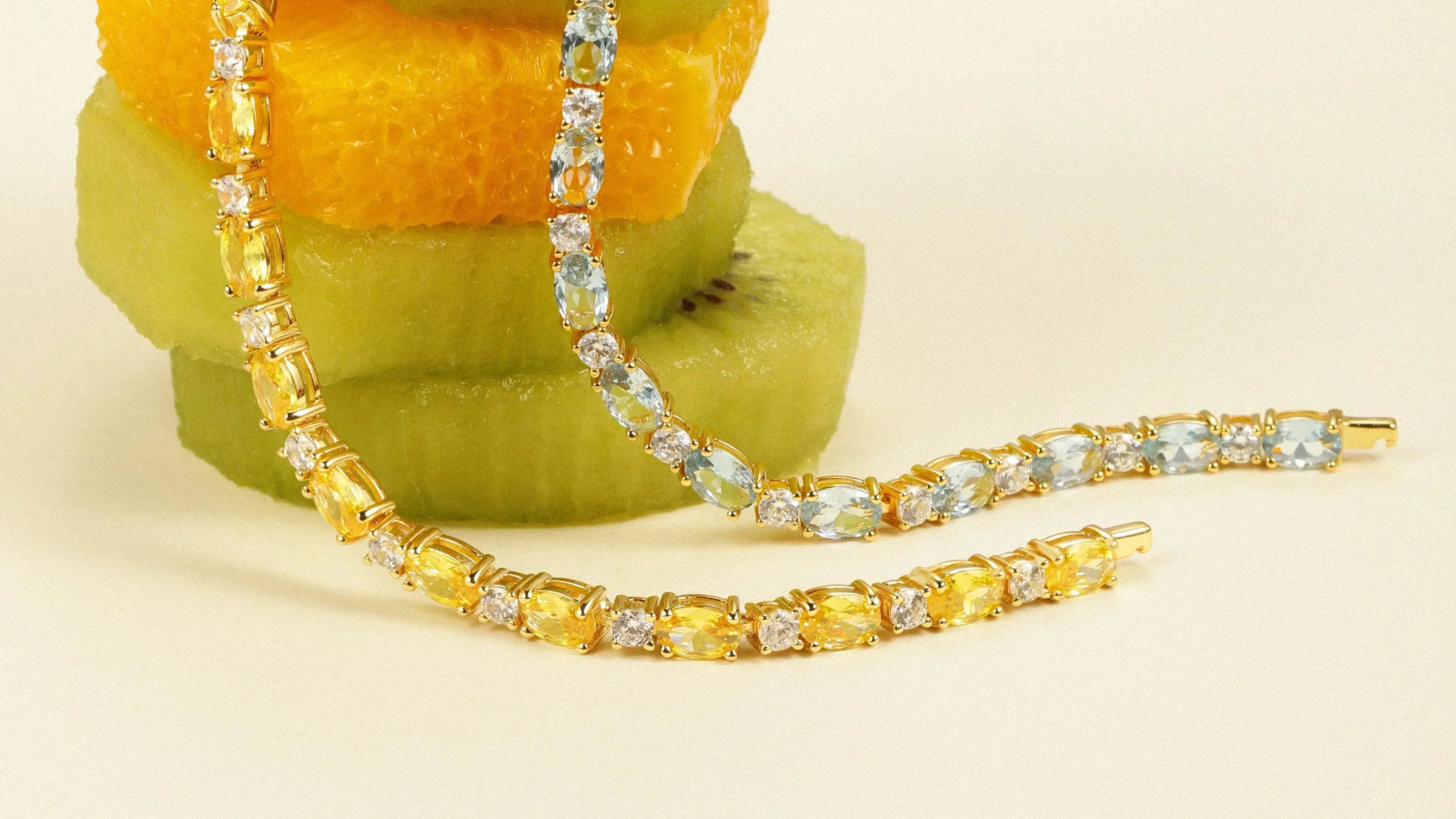 Two gemstone bracelets, one with yellow stones and one with blue stones, draped over orange and kiwi slices.